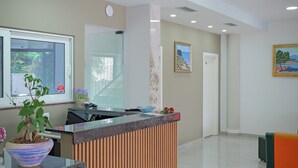 A reception area with a wooden reception desk, a potted plant, a window with a view, and a painting on the wall. - Hotel Kroi (Ksamil)
