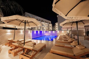 A poolside area with lounge chairs and umbrellas, a pool, and a building in the background. - Hotel Kroi (Ksamil)