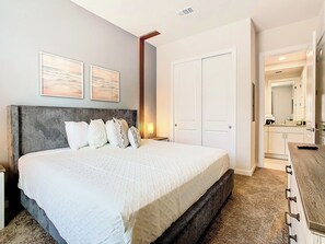 A hotel room with a large bed, a bedside table, a nightstand, a wall-mounted lamp, a door, and a view into the adjacent bathroom.