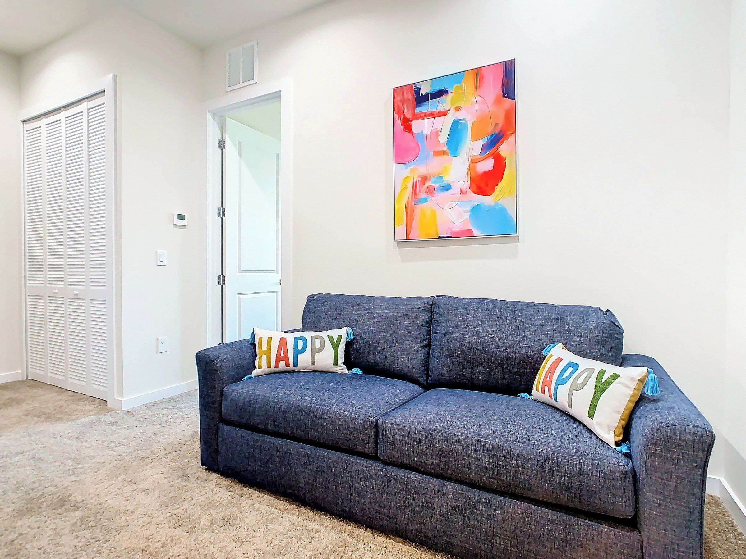A room with a dark grey sofa, two pillows with 'HAPPY' text, a colorful abstract painting on the wall, a white door, and a beige carpet.