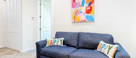 Une pièce avec un canapé gris foncé, deux coussins ornés du mot « HAPPY », un tableau abstrait aux couleurs vives accroché au mur, une porte blanche et un tapis beige.