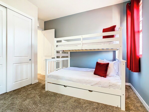 Una cama litera con una cama inferior y una individual, ropa de cama blanca, una almohada roja, una puerta y suelo alfombrado.