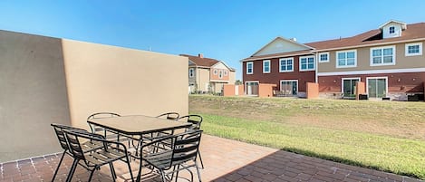 Un patio con mesa y sillas, un sendero de ladrillo y un edificio con varias ventanas y una puerta.