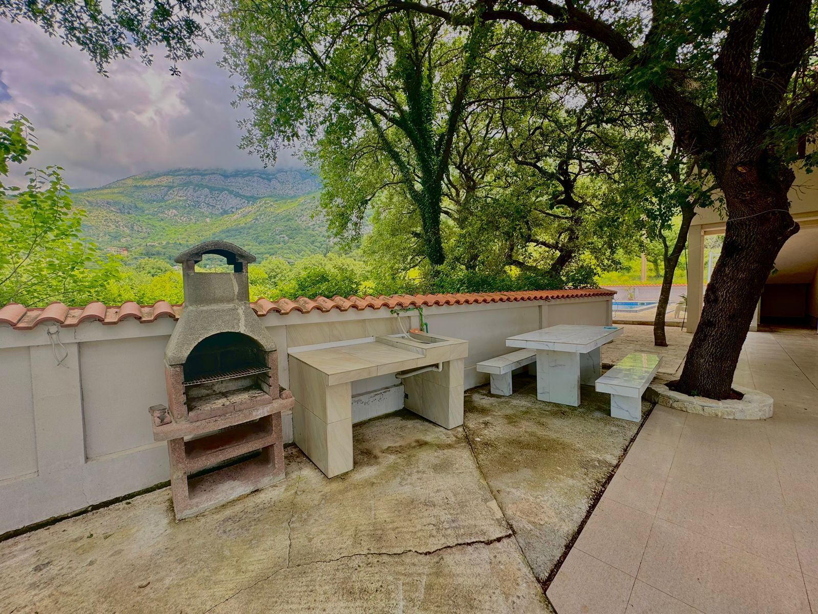 Een terras buiten met een stenen barbecue, een spoelbak en een tafel, omringd door bomen en uitzicht op een glooiend landschap.