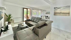 A modern living room with a grey sectional sofa, a small coffee table, and a potted plant. There is a sliding glass door leading to an outdoor area with a table and chairs. - Stars Residence (Petrovac)
