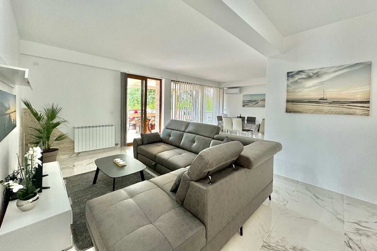 A modern living room with a grey sectional sofa, a small coffee table, and a potted plant. There is a sliding glass door leading to an outdoor area with a table and chairs.