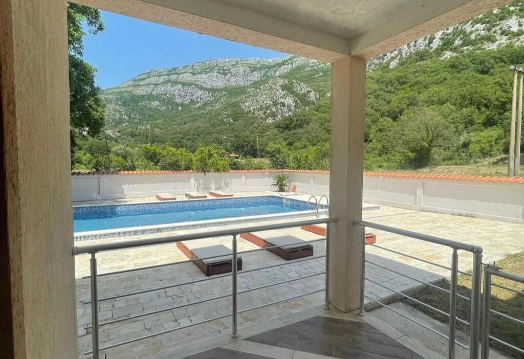 A covered outdoor area with a swimming pool, lounge chairs, and a view of a mountainous landscape.