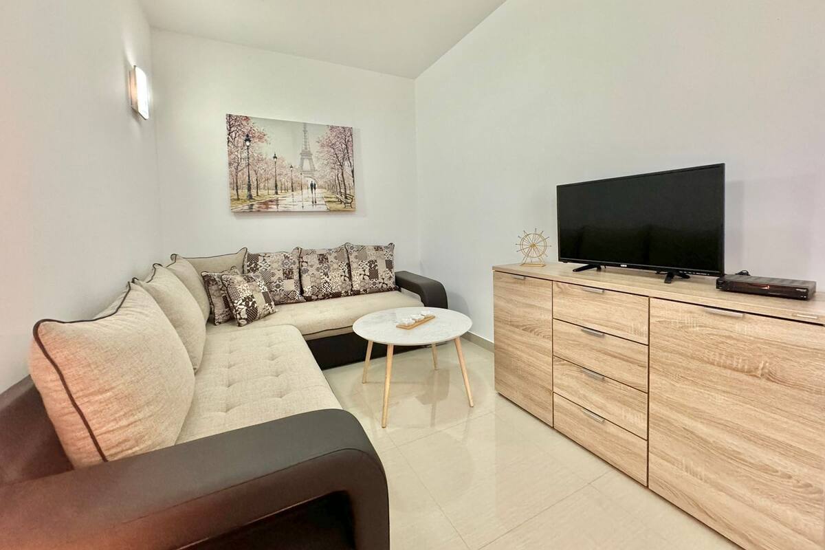 A modern living room with a sofa, a coffee table, a TV on a wooden cabinet, and a painting of a park scene.
