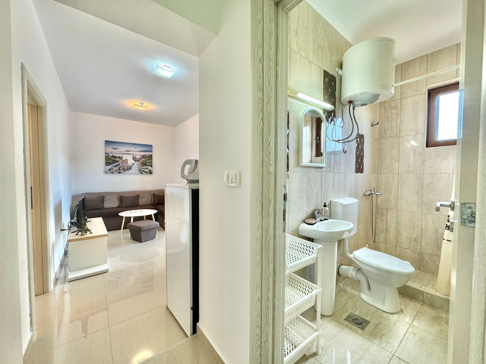 Un baño moderno con inodoro, lavamanos y ducha. En el área de estar contigua hay una pequeña mesa y un sofá.