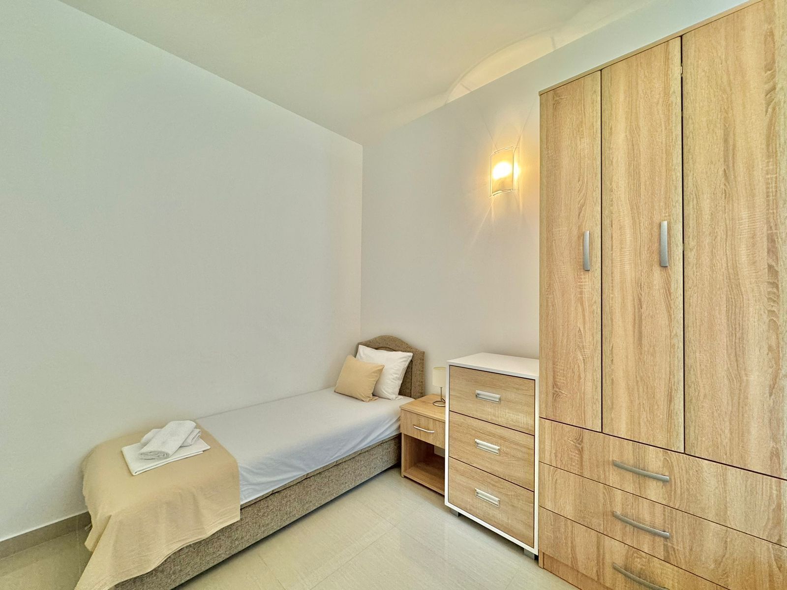 A small, neatly arranged bedroom with a single bed, a bedside table, and a wooden wardrobe with multiple drawers.