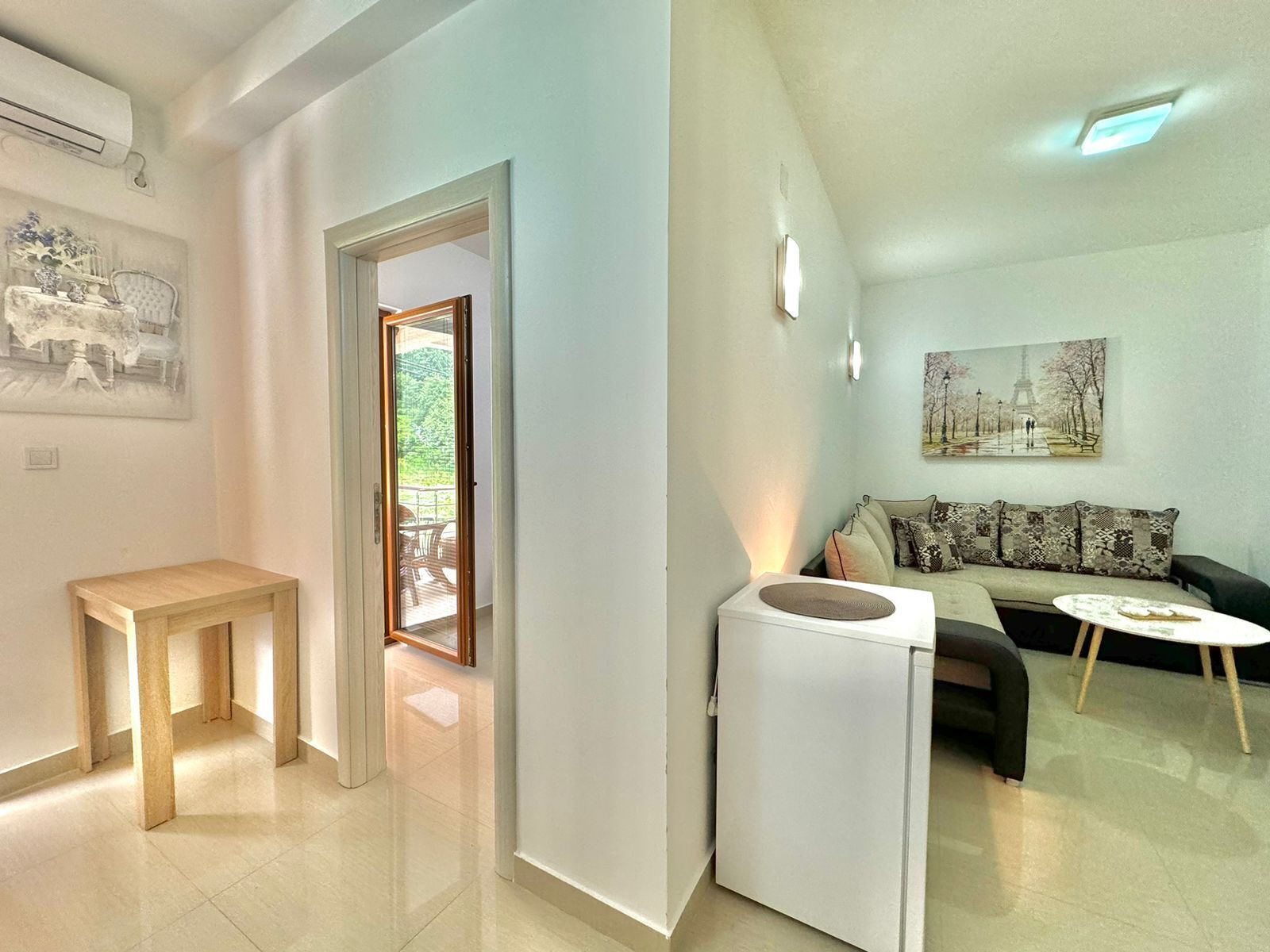 A modern living room with a sofa, a coffee table, and a small white table. There is a door leading to a balcony with outdoor furniture.