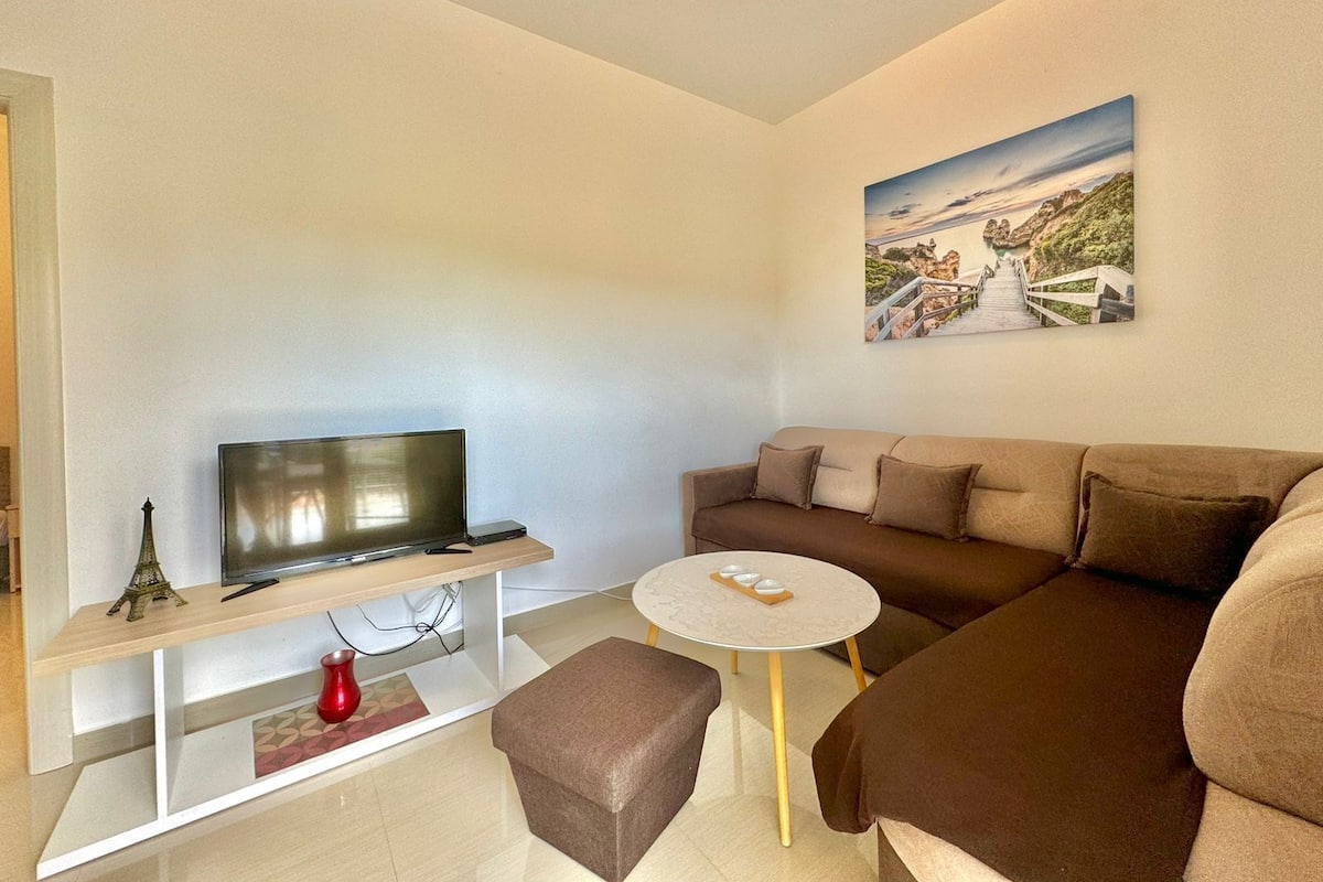 A modern living room with a flat-screen TV on a white stand, a brown sofa, a small round table with a white top, and a painting of a coastal landscape on the wall.