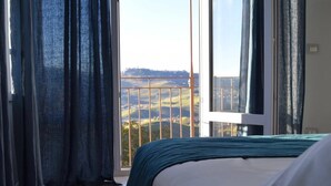 Une chambre à coucher avec un lit, une vue sur la campagne par une grande fenêtre et un balcon avec une balustrade.