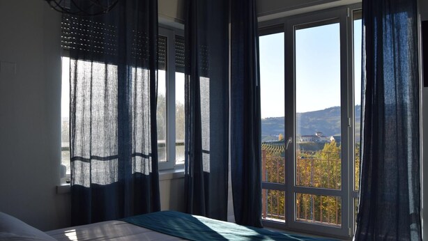 Une chambre à coucher avec un lit, une grande fenêtre avec des rideaux et une vue sur un paysage composé d’arbres et de collines.