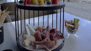 Une table surmontée d’un présentoir à trois niveaux où sont disposés divers aliments, une bougie et un verre d’eau, le tout placé devant un décor représentant une plage et un ciel nuageux.
