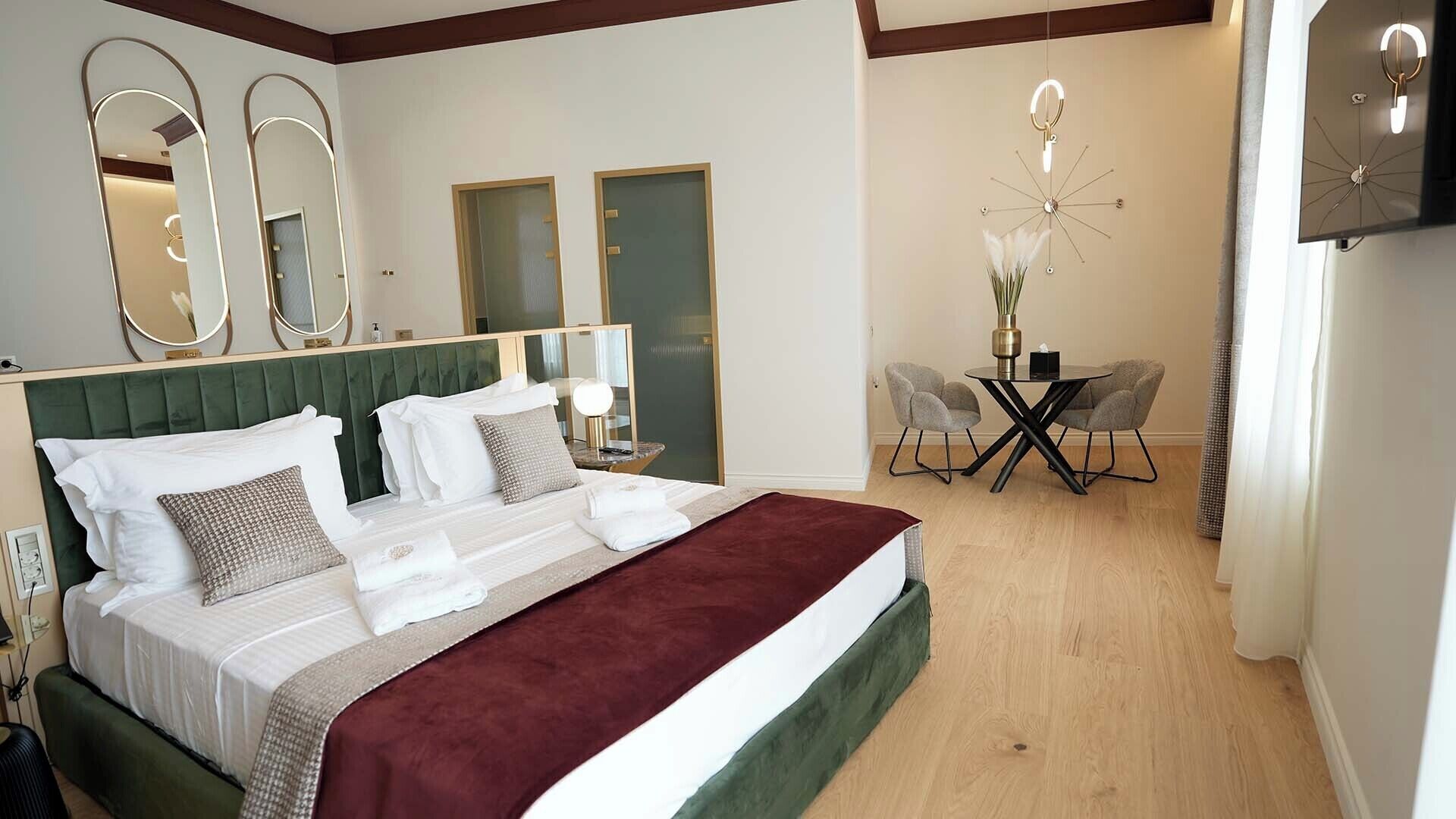 A hotel room with a large bed, two doors, a dining area with a table and chairs, and a TV mounted on the wall.
