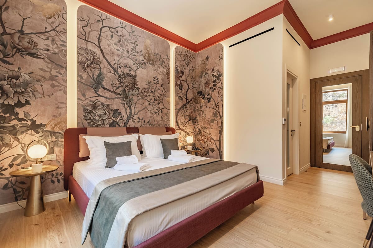 A bedroom with a large bed, a nightstand with a lamp, and a wall with floral wallpaper.