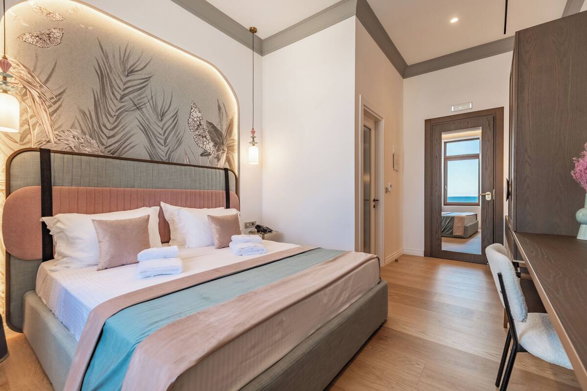 A modern bedroom with a large bed, a desk, and a view of the sea through an open door.