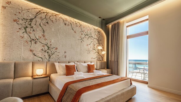 Une chambre d’hôtel moderne dotée d’un grand lit, d’un canapé, d’une fresque murale et d’un balcon offrant une vue sur la mer.