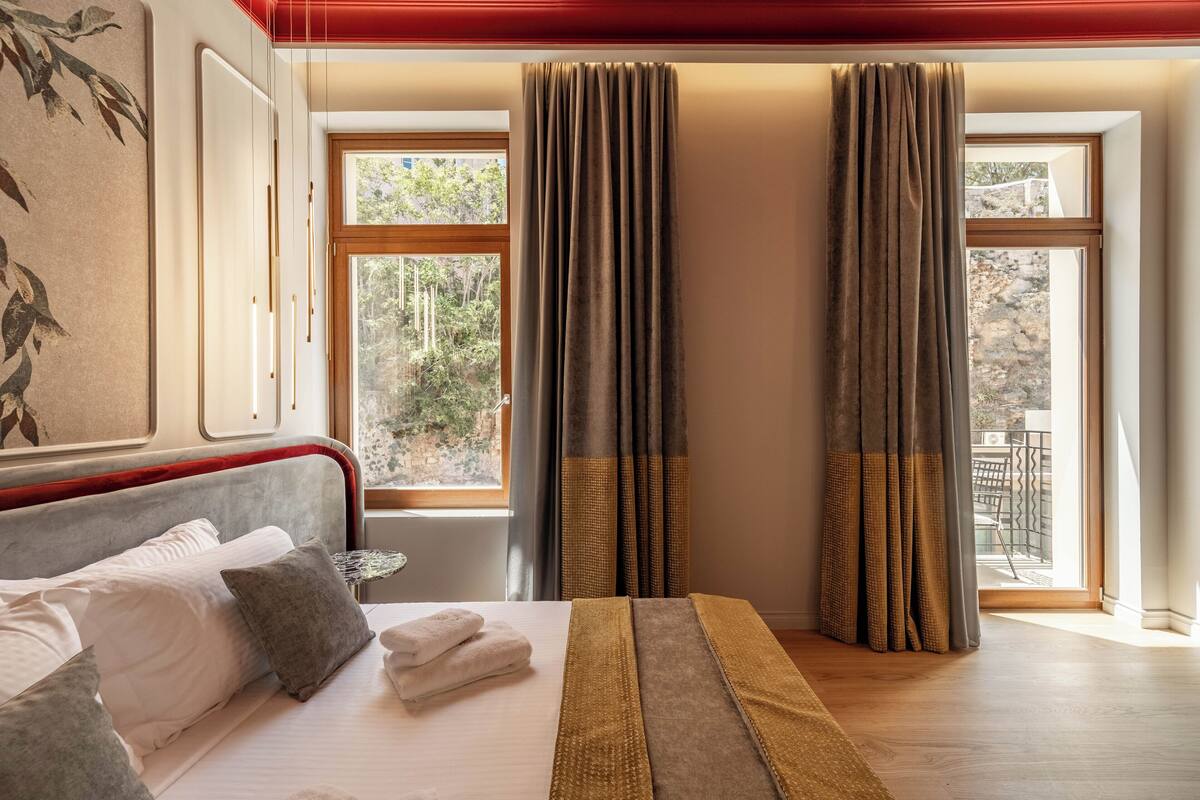 A neatly made bed with pillows and a blanket, a window with curtains, and a view of greenery outside.
