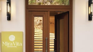 Une porte à deux battants en bois, avec un panneau de verre, une poignée et une sonnette. Un panneau indiquant « Mira Vita Boutique Hotel » est fixé au mur, sur la gauche.