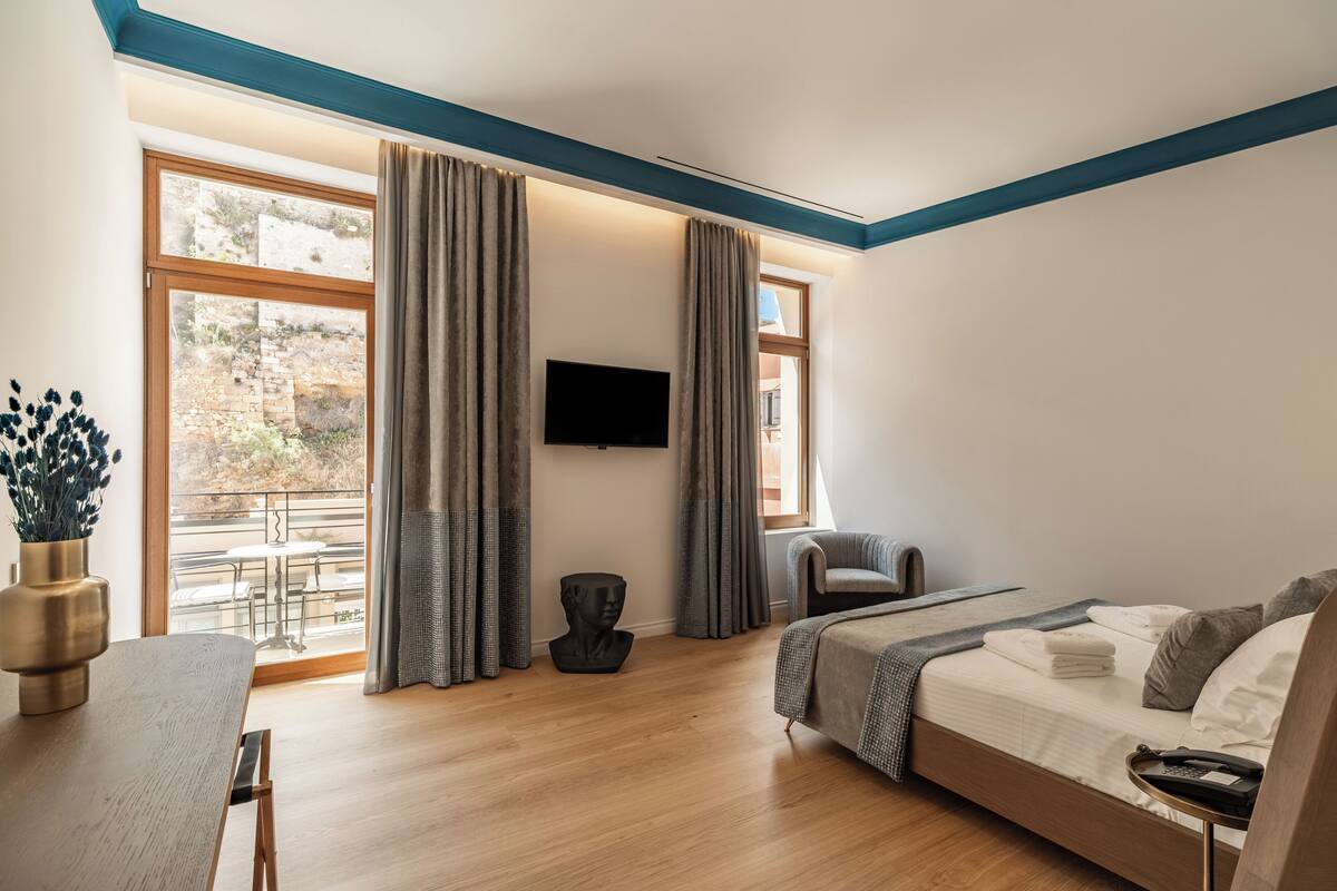 A modern hotel room with a large window, a flat-screen TV, a bed, a desk, and a chair.