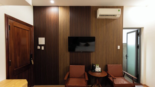 A hotel room with a wooden panel wall, a flat-screen TV mounted above a small table with two leather chairs, and a door leading to another room. - LuckyStar Hotel (Pleiku)