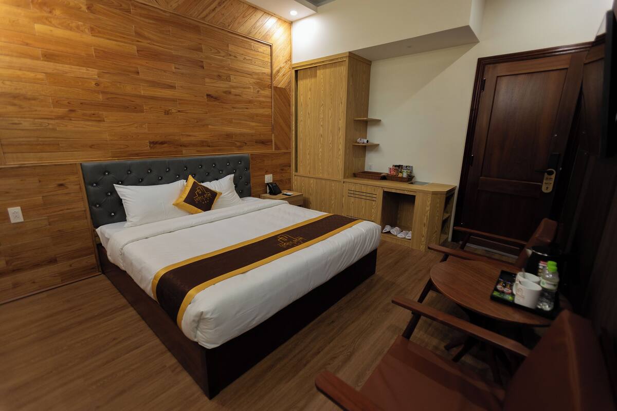 A hotel room with a large bed, wooden panel walls, a wooden wardrobe, a small wooden table with a tray, and a door with a handle.