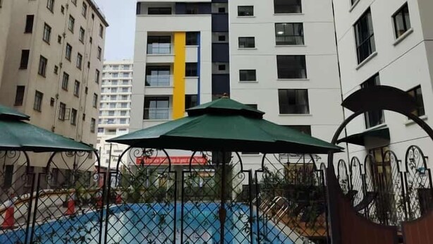 A high-rise building with a yellow vertical stripe, surrounded by other buildings. There are outdoor tables and chairs with green umbrellas, and a swimming pool in the background.