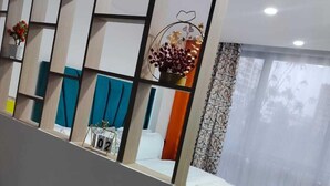 A hotel room with a bed, a window with sheer curtains, a decorative wall panel, and a small table with a vase.