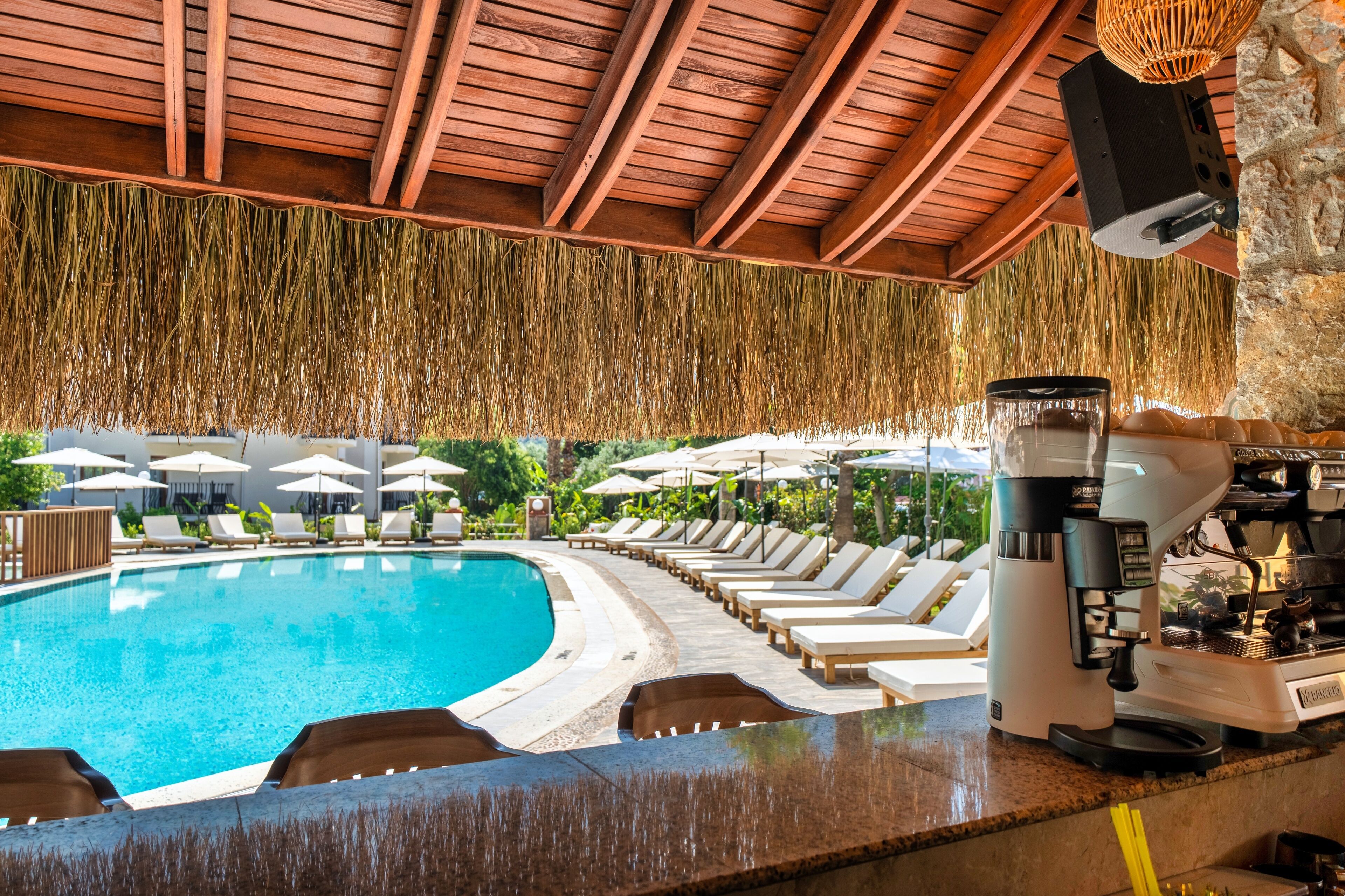 A poolside area with lounge chairs and umbrellas, a coffee machine, and a stone wall.