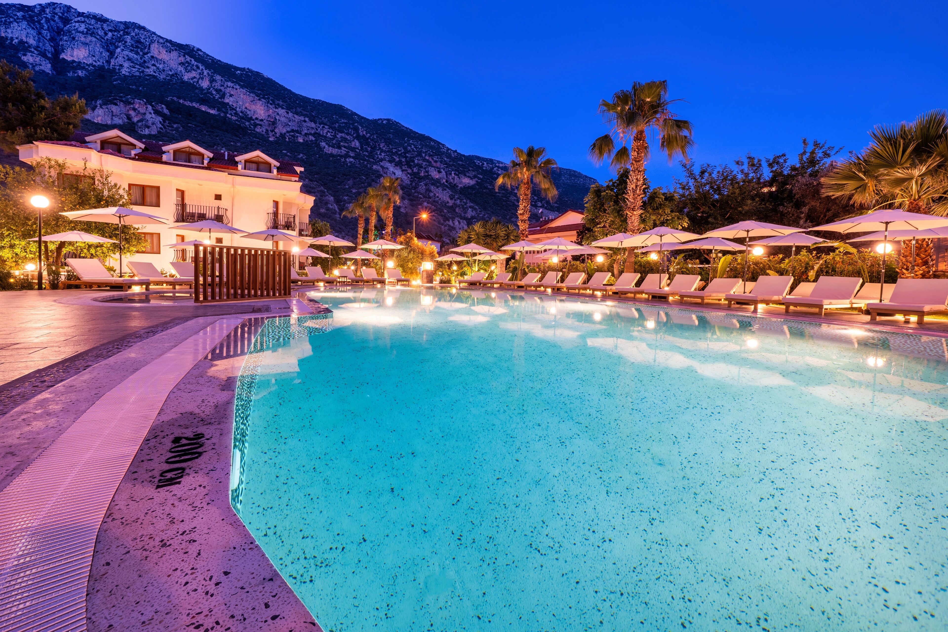 A swimming pool with lounge chairs and umbrellas, surrounded by a building with balconies and palm trees, set against a mountainous backdrop.