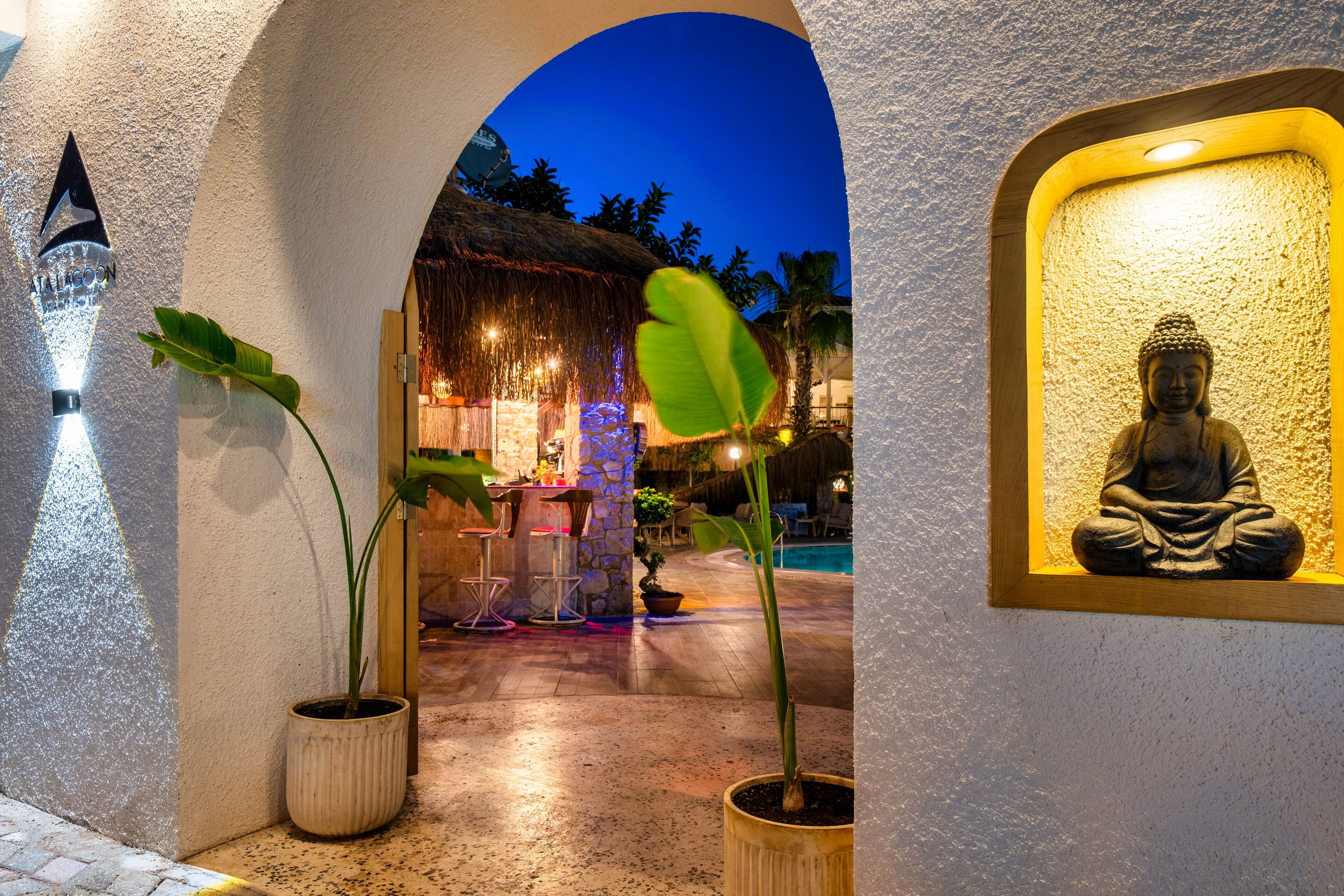 A well-lit outdoor area with a Buddha statue, potted plants, and a thatched-roof structure.