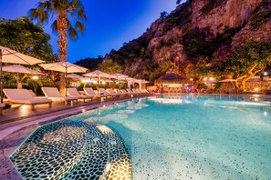 Zona de piscina con sillones reclinables y sombrillas, rodeada por un paisaje montañoso.