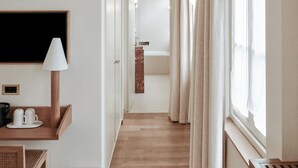 Un couloir d’hôtel avec un plancher en bois, un bureau avec une lampe et un lit visible au loin.