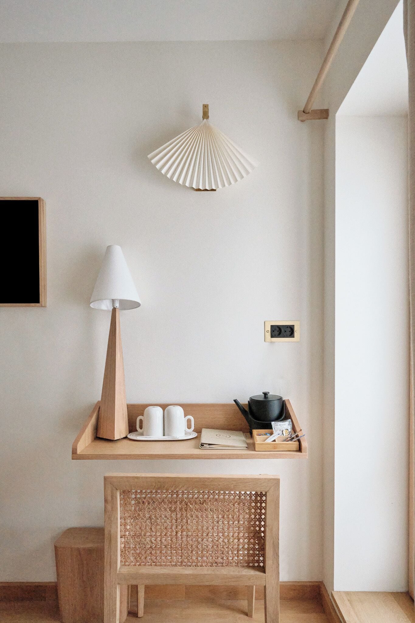 Una habitación minimalista con un escritorio de madera, una silla tejida, una lámpara blanca y un estante de pared con tazas y una tetera.