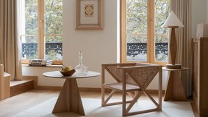 Une pièce avec une table ronde en bois, une chaise en osier, un lampadaire et une fenêtre donnant sur des arbres.