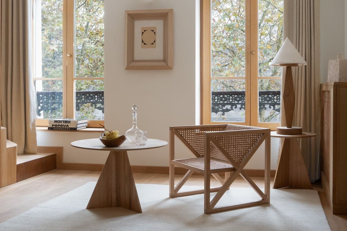 Una habitación con una mesa redonda de madera, una silla de mimbre, una lámpara de pie y un ventanal con vistas a árboles.