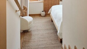 Un couloir étroit mène à une chambre qui contient un lit, une commode, une fenêtre avec des rideaux et un petit tabouret.
