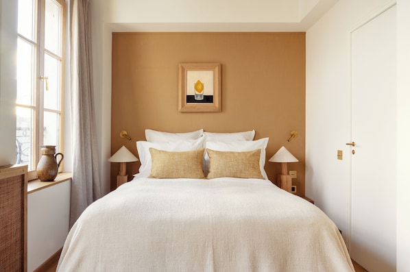 A neatly made bed with white and beige bedding, a framed picture on the wall, and a window with curtains. - Hôtel Elysée Montmartre (Paris)