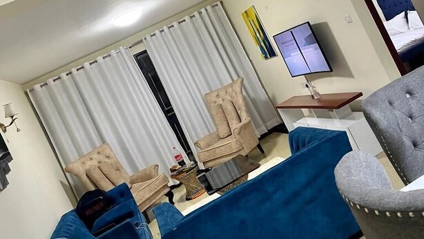 Una sala de estar con un sofá azul, un sillón beige y otro sillón gris. Hay un televisor sobre un mueble bajo y una ventana con cortinas.