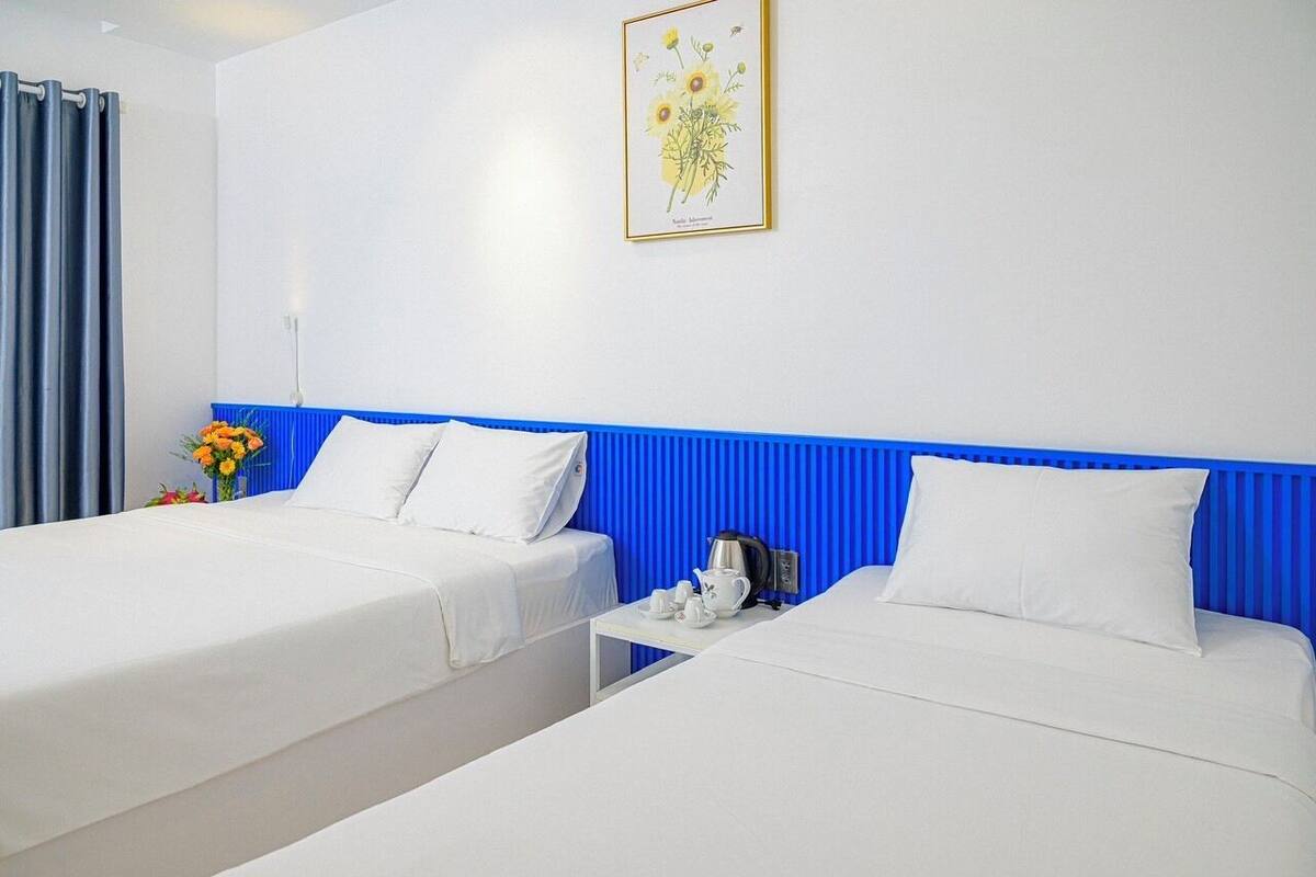 Une chambre d’hôtel avec deux lits, une tête de lit bleue, une œuvre d’art florale encadrée et une petite table avec une bouilloire et des tasses.
