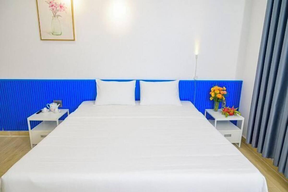 Une chambre d’hôtel avec un grand lit, une tête de lit bleue, du linge de lit blanc, deux tables de chevet avec des lampes et un tableau encadré au mur.