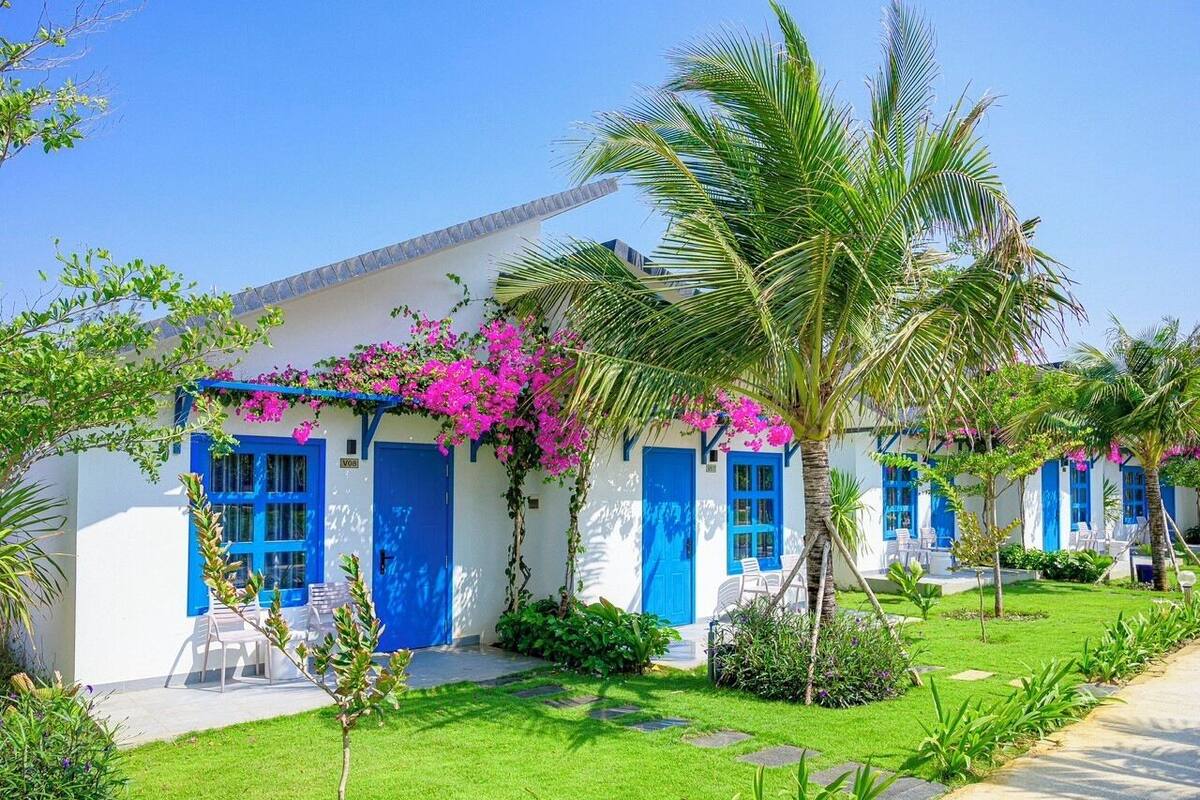 Un bâtiment blanc aux portes et fenêtres bleues, entouré d’une végétation luxuriante et de fleurs roses.