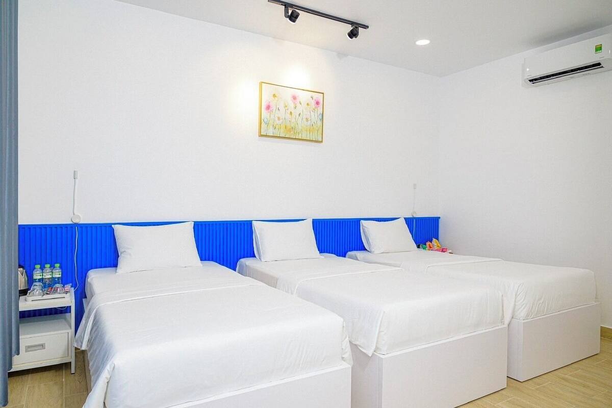 Une chambre d’hôtel avec deux lits simples, une tête de lit bleue, du linge de lit blanc, une petite table de chevet avec des articles de toilette et un tableau au mur.