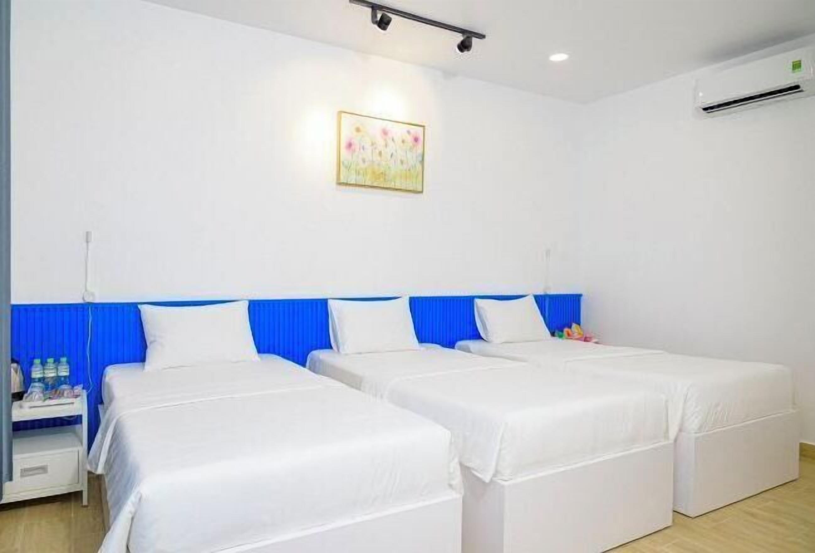 A hotel room with two single beds, a blue headboard, white bedding, a small bedside table with water bottles, and a wall-mounted air conditioner.