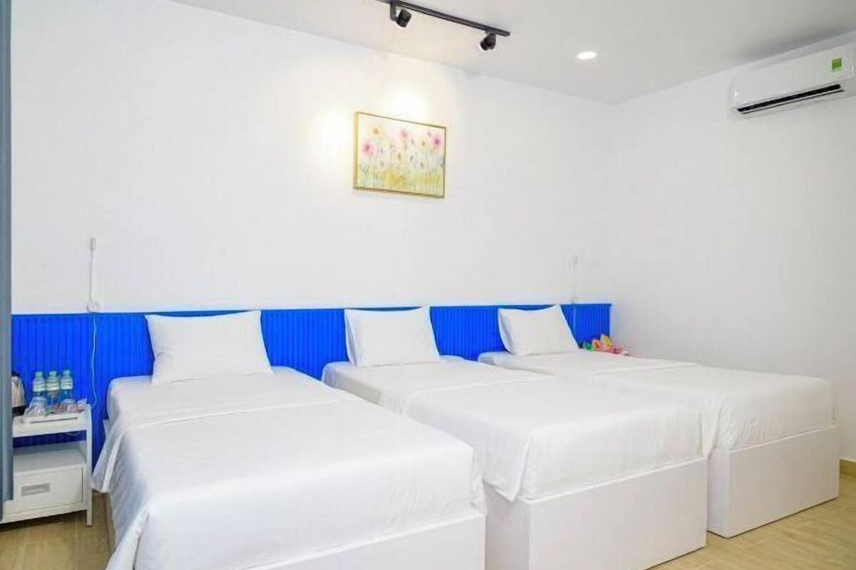 Une chambre d’hôtel avec deux lits simples, une tête de lit bleue, du linge de lit blanc, une petite table de chevet avec des bouteilles d’eau et un climatiseur fixé au mur.