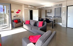 Un salon moderne avec un canapé d’angle gris, des coussins rouges, un coin repas avec une table et des chaises, une cuisine avec des placards et un plan de travail, et une porte-fenêtre coulissante donnant sur un balcon.