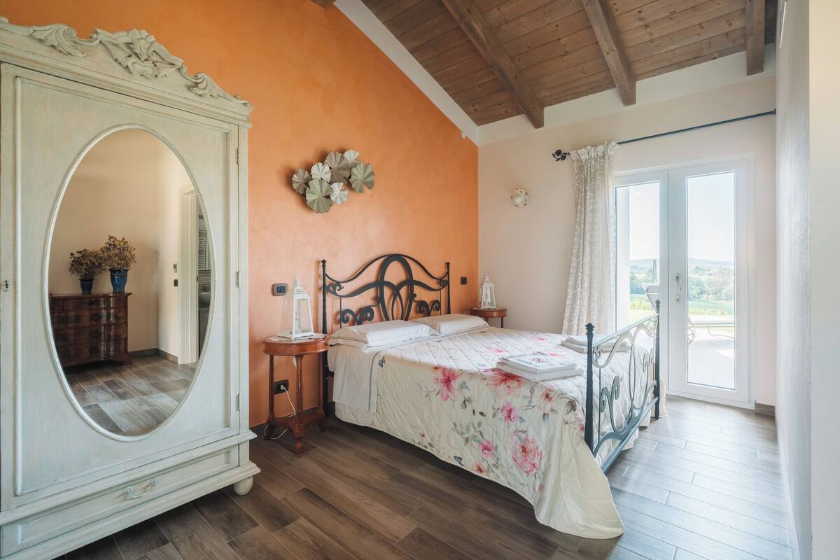 A bedroom with a large mirror, a bed with floral bedding, a wooden dresser, and a view of the outdoors through a sliding door.