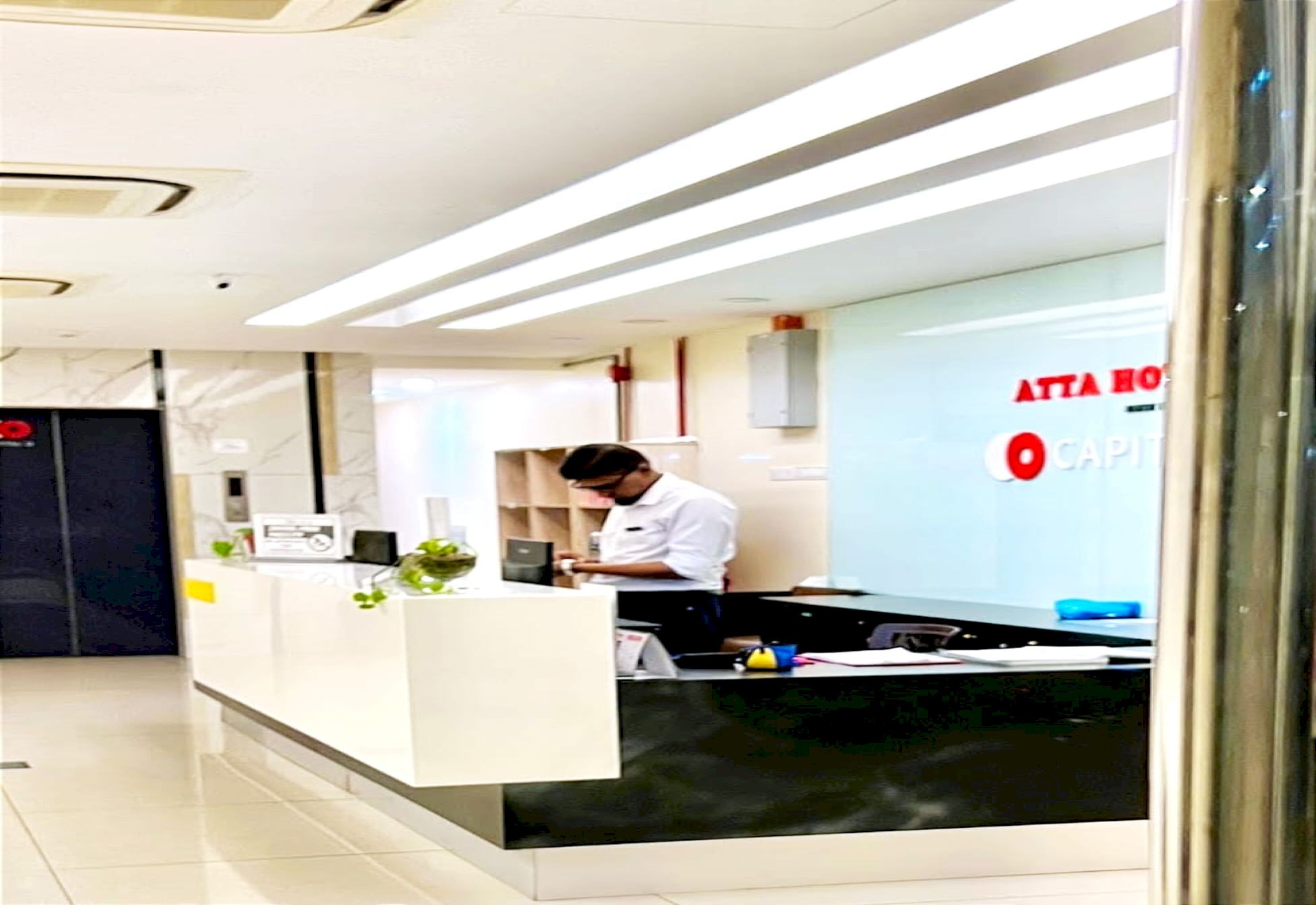 A reception desk with a man working behind it, a sign reading "ATTA HOUSE CAPITOL," and a modern interior design.